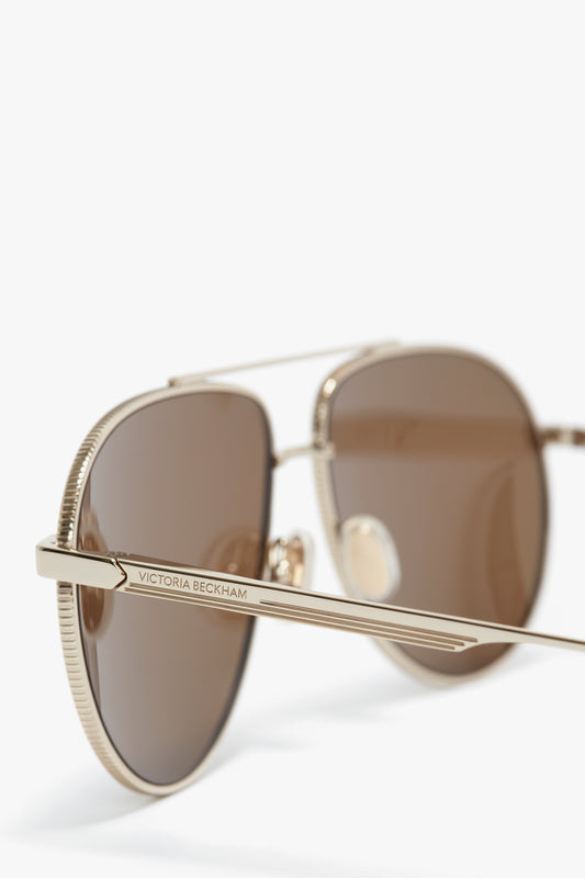 Close-up of a pair of Victoria Beckham V Metal Pilot Sunglasses In Gold-Khaki with brown lenses and gold metal frames, featuring the Victoria Beckham logo on the temple and adjustable nose pads for a perfect fit.