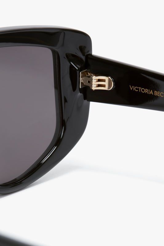 Close-up of black oversized frames with dark lenses, featuring "VICTORIA BECKHAM" in gold text on the arm. The image focuses on the hinge area of the frame, showcasing the impeccable design of Victoria Beckham Acetate Visor Sunglasses In Black.