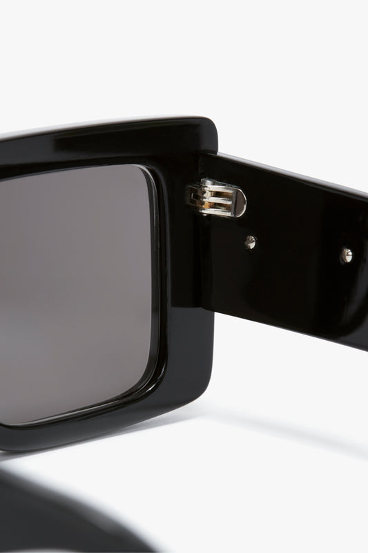 Close-up of black oversized sunglasses frame with a visible hinge connecting the temple arm to the front of the bold, rectangular lenses. The Victoria Beckham signature Oversized Frame Sunglasses In Black adds a touch of distinctive style.