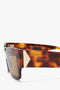 Close-up of V Plaque Rectangular Frame Sunglasses In Tortoise with thick frames, gold-tone V plaque accents, and "Victoria Beckham" inscribed on the arm.