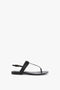 The **Victoria Beckham Flat Chain Sandal In Black Leather** is a black thong sandal with a flat sole, crafted from luxurious calfskin leather and featuring an adjustable ankle strap adorned with a signature chain detail.