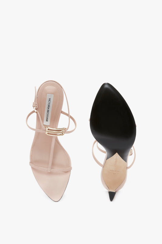 A pair of Victoria Beckham Frame Detail Sandal In Nude Leather high-heeled sandals with nude straps and gold buckles. One shoe, featuring a sculptural heel, is shown from the top view, while the other from the bottom view displays the black sole and pointed toe with an adjustable ankle strap.