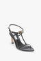 A Frame Detail Sandal In Black Leather by Victoria Beckham with thin straps and a gold-tone B frame buckle, featuring a V-shaped sculptural heel.