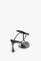 A Victoria Beckham Frame Detail Sandal In Black Leather featuring a thin ankle strap and a V-shaped sculptural heel, crafted from supple nappa leather, set against a white background.