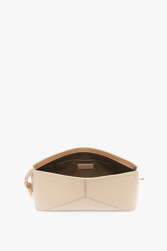 A beige shoulder bag with an open top, revealing a spacious brown interior and a small inner pocket with a beige label. The Victoria Clutch Bag In Sesame Leather, crafted for Victoria Beckham's brand SS24 runway collection, features gold hardware and a geometric design on the exterior, reminiscent of their iconic calf leather clutch.