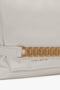 Close-up image of a Victoria Beckham handbag, specifically the Puffy Chain Pouch With Strap In White Leather, featuring a white design with gold chain detail and "VICTORIA BECKHAM" elegantly printed in gold letters on the leather.