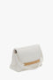 A white Victoria Beckham handbag with a rectangular shape, a fold-over flap, and a gold-tone chain accent. The Puffy Chain Pouch With Strap In White Leather features an adjustable strap and is photographed against a plain white background.