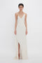 A person stands against a white backdrop wearing an Exclusive Asymmetric Bias Frill Dress In Ivory by Victoria Beckham, with thin straps and diagonal ruffle details, crafted from luxurious crepe back satin.