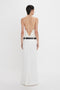 A person with short dark hair is wearing a backless white dress with a thin black belt and an Exclusive Tailored Floor-Length Pleated Skirt In Ivory by Victoria Beckham, standing and facing away from the camera against a plain white background.