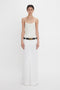 A person stands against a white background, wearing an Exclusive Cami Top In Ivory by Victoria Beckham and a long white skirt with a black belt featuring a gold buckle.