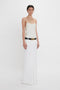 A person stands against a plain background, wearing a sleeveless Exclusive Cami Top In Ivory by Victoria Beckham and a long white skirt with a black belt.