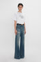 Person wearing a white T-shirt with the text "DAVID'S WIFE" and blue high-waisted wide-leg jeans, posed against a plain white background. The seventies-style outfit is completed with vintage denim reminiscent of Victoria Beckham’s Alina High Waisted Jean In Indigrey Wash.