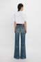 A person stands facing away, wearing a white T-shirt and the Alina High Waisted Jean In Indigrey Wash by Victoria Beckham with a leather belt.