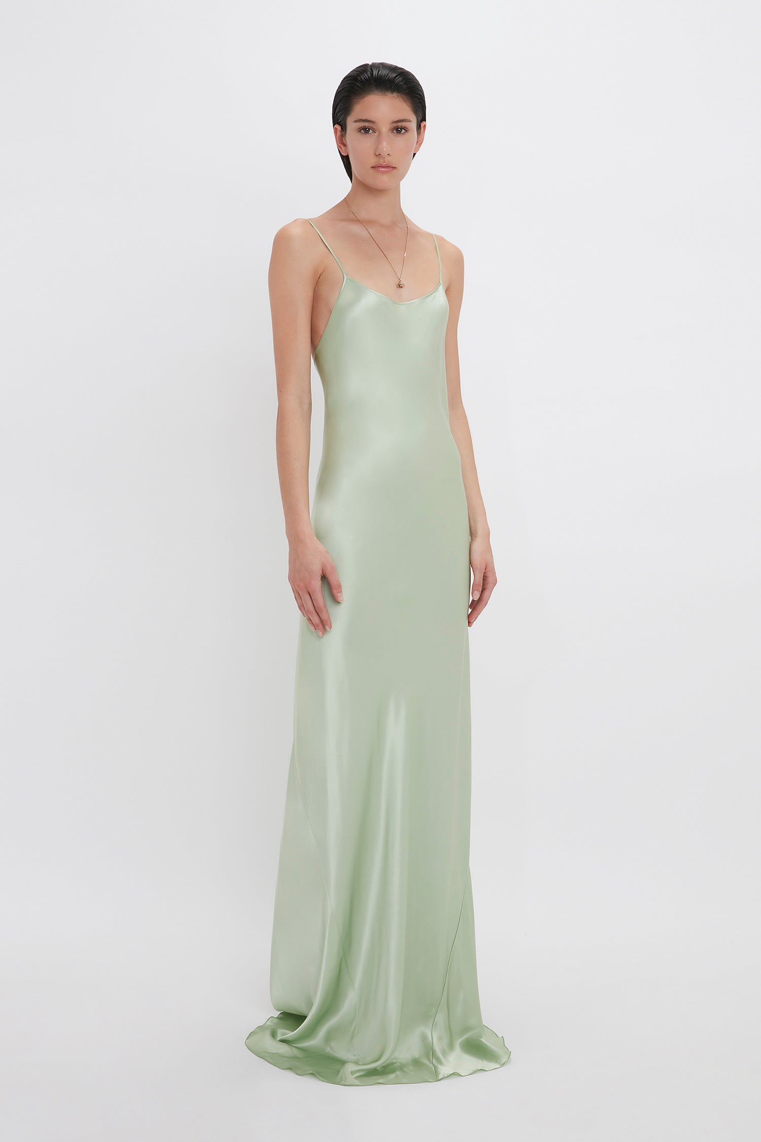 Person standing against a white background wearing a floor-length, satin, light green dress with thin spaghetti straps and a small Exclusive Camellia Flower Necklace In Gold by Victoria Beckham handcrafted in Italy.