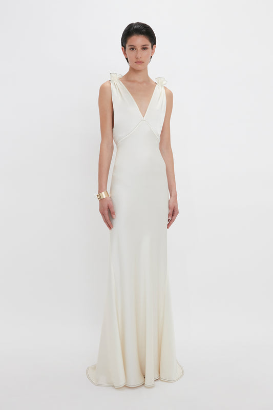 A person stands facing forward, wearing a sleeveless, floor-length white Gathered Shoulder Floor-Length Cami Gown In Ivory by Victoria Beckham made of crepe back satin with a V-neckline and slight train, and a gold bracelet on the left wrist. The background is plain white.