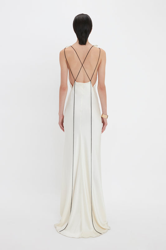 A person is standing with their back to the camera, wearing a Gathered Shoulder Floor-Length Cami Gown In Ivory with thin black stripes and crisscross straps by Victoria Beckham. The backless gown gracefully extends to the floor.