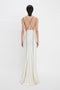 A person is standing with their back to the camera, wearing a Gathered Shoulder Floor-Length Cami Gown In Ivory with thin black stripes and crisscross straps by Victoria Beckham. The backless gown gracefully extends to the floor.