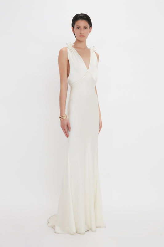 A person is standing and wearing a long, white, sleeveless "Gathered Shoulder Floor-Length Cami Gown In Ivory" by Victoria Beckham with a deep V-neckline and gathered shoulders. They have short dark hair and are accessorized with bracelets on one wrist.