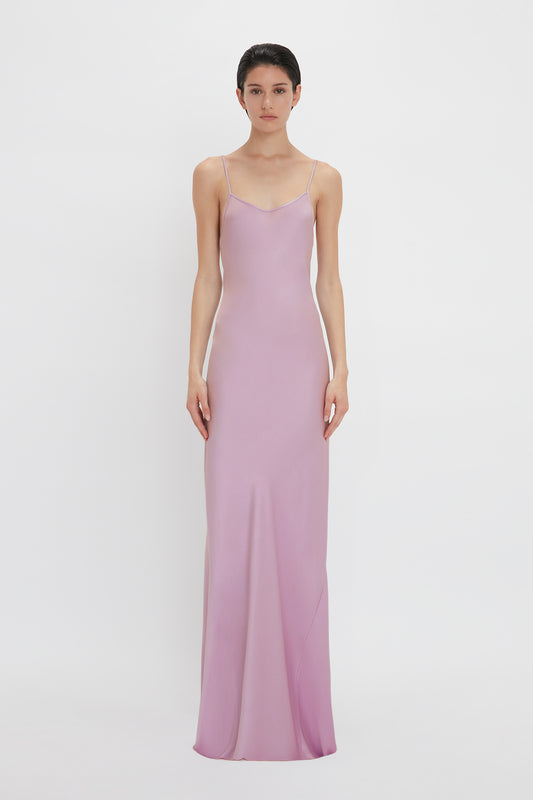 A person is standing against a white background wearing a sleeveless, floor-length, form-fitting Victoria Beckham Low Back Cami Floor-Length Dress In Rosa with thin straps.