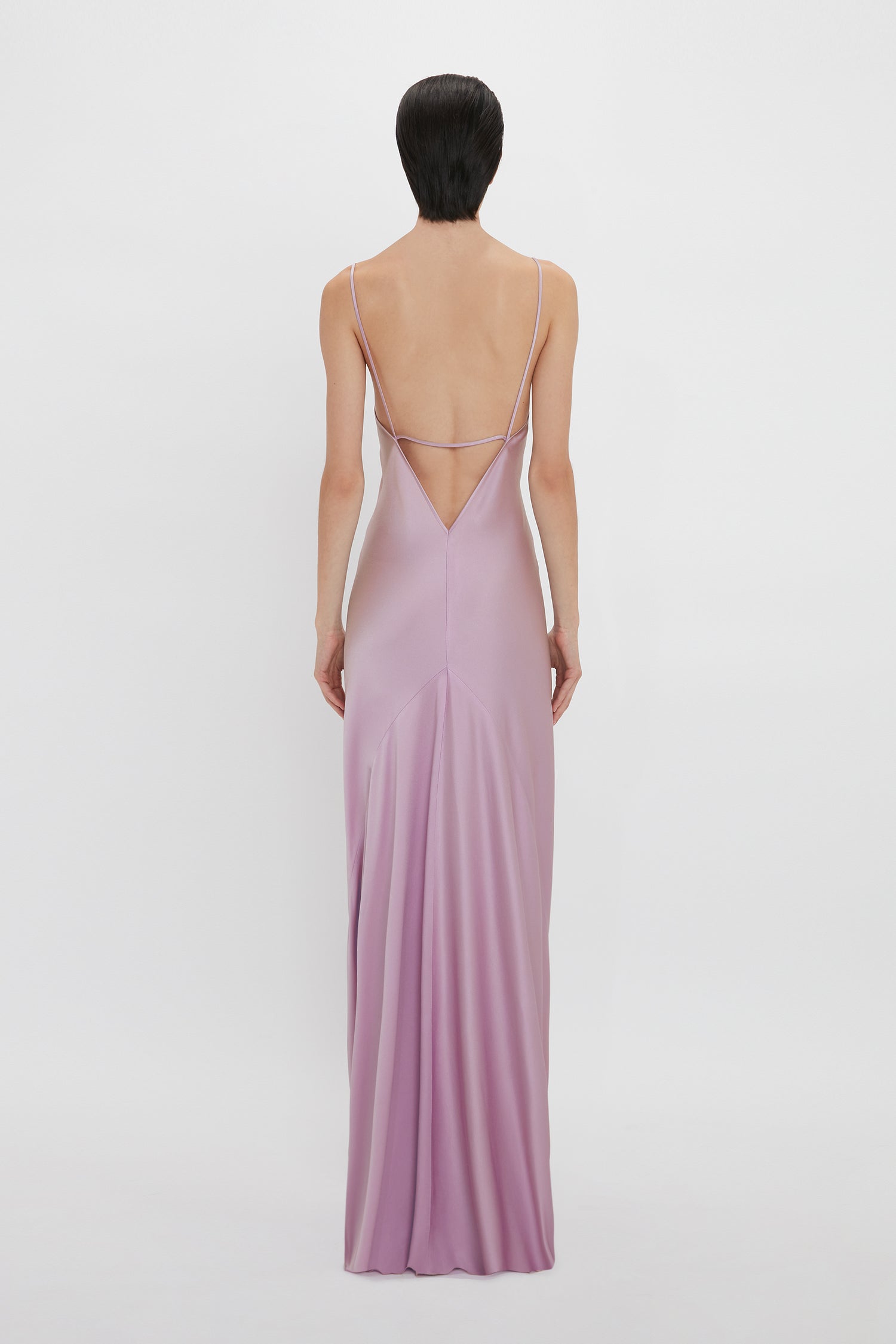 A woman with short, dark hair is wearing a backless, floor-length lilac satin slip dress with thin straps, reminiscent of Victoria Beckham's elegant designs. She is shown from the back in the Low Back Cami Floor-Length Dress In Rosa by Victoria Beckham.