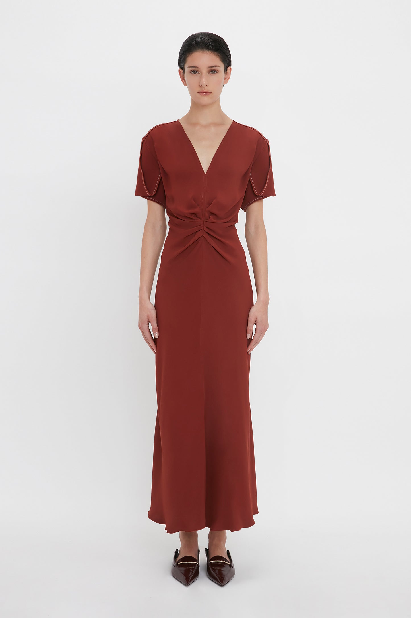 A person stands against a white background wearing a russet Gathered V-Neck Midi Dress In Russet by Victoria Beckham with a cinched waist and short sleeves, complemented by pointed dark brown shoes.