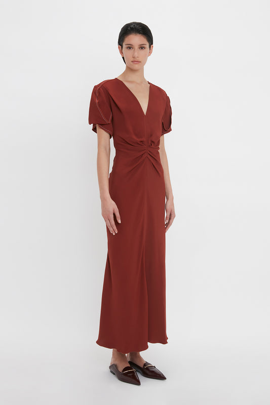 A person is standing against a plain white background, wearing a russet, floor-length dress with short sleeves and brown flat shoes. The Gathered V-Neck Midi Dress In Russet by Victoria Beckham features a gathered v-neck design in figure-flattering stretch fabric.