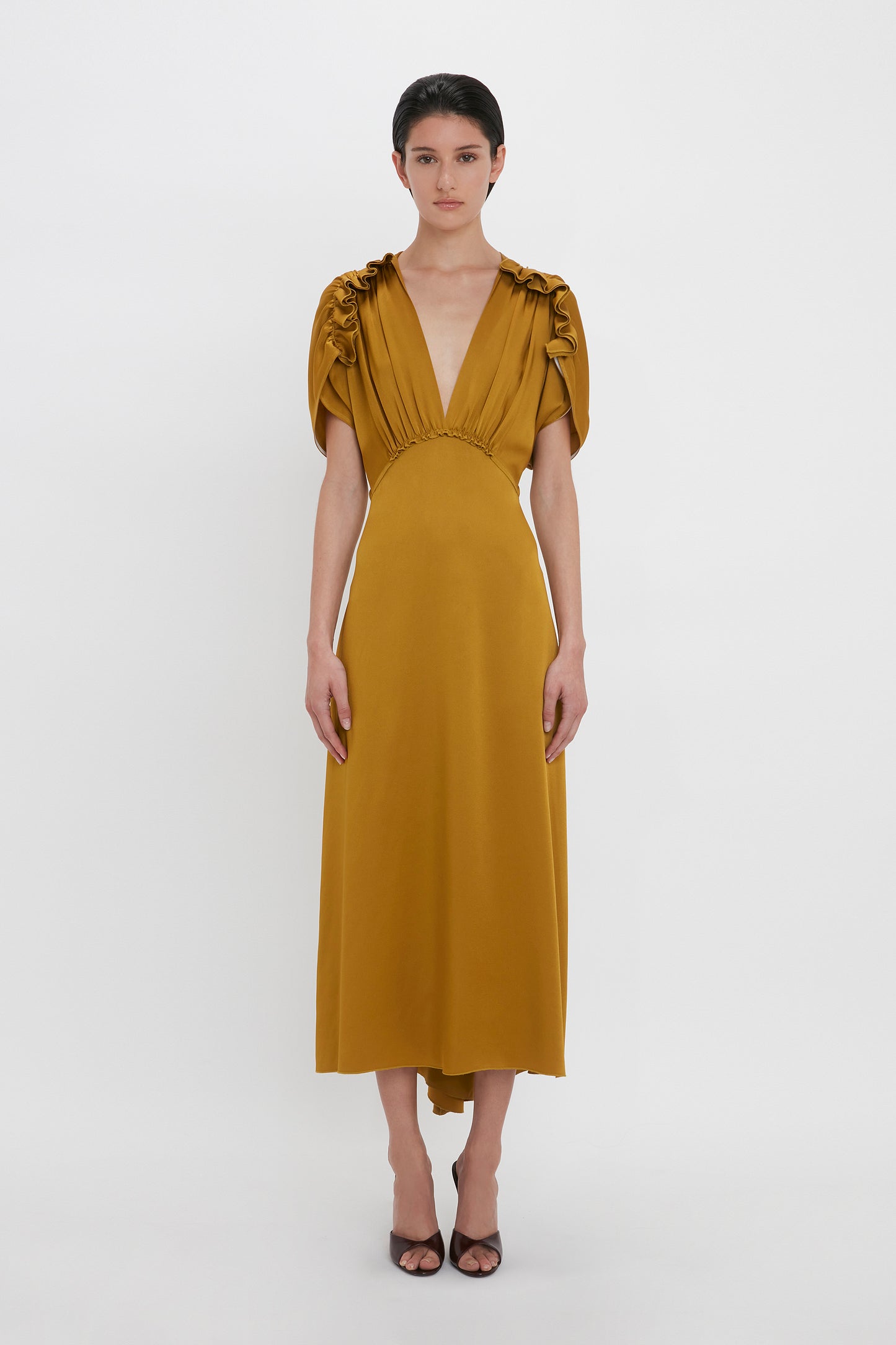 A person stands against a plain white background, wearing a Victoria Beckham V-Neck Ruffle Midi Dress In Harvest Gold with gathered shoulder details. Exuding a seductive quality, they are also wearing black open-toed heels.