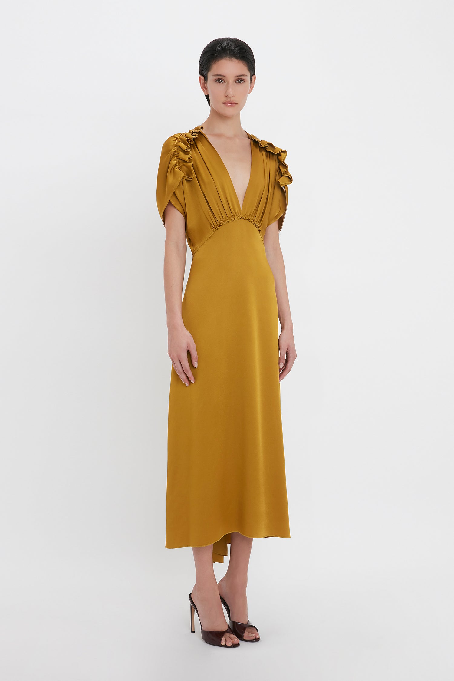 A person stands in a Victoria Beckham V-Neck Ruffle Midi Dress In Harvest Gold with puffed sleeves, paired with black heeled shoes, exuding a seductive quality against a plain white background.