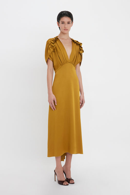 A person stands in a Victoria Beckham V-Neck Ruffle Midi Dress In Harvest Gold with puffed sleeves, paired with black heeled shoes, exuding a seductive quality against a plain white background.