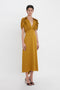 A person stands in a Victoria Beckham V-Neck Ruffle Midi Dress In Harvest Gold with puffed sleeves, paired with black heeled shoes, exuding a seductive quality against a plain white background.