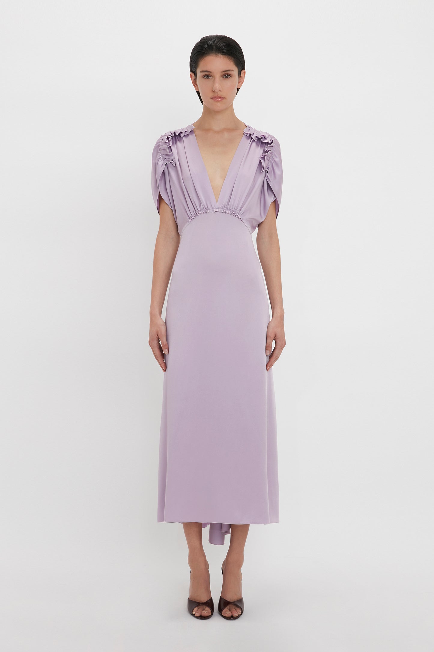 A person wearing a light purple, Victoria Beckham V-Neck Ruffle Midi Dress In Petunia stands against a plain white background. They have short dark hair and are sporting dark open-toe shoes.