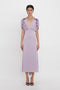 A person wearing a light purple, Victoria Beckham V-Neck Ruffle Midi Dress In Petunia stands against a plain white background. They have short dark hair and are sporting dark open-toe shoes.