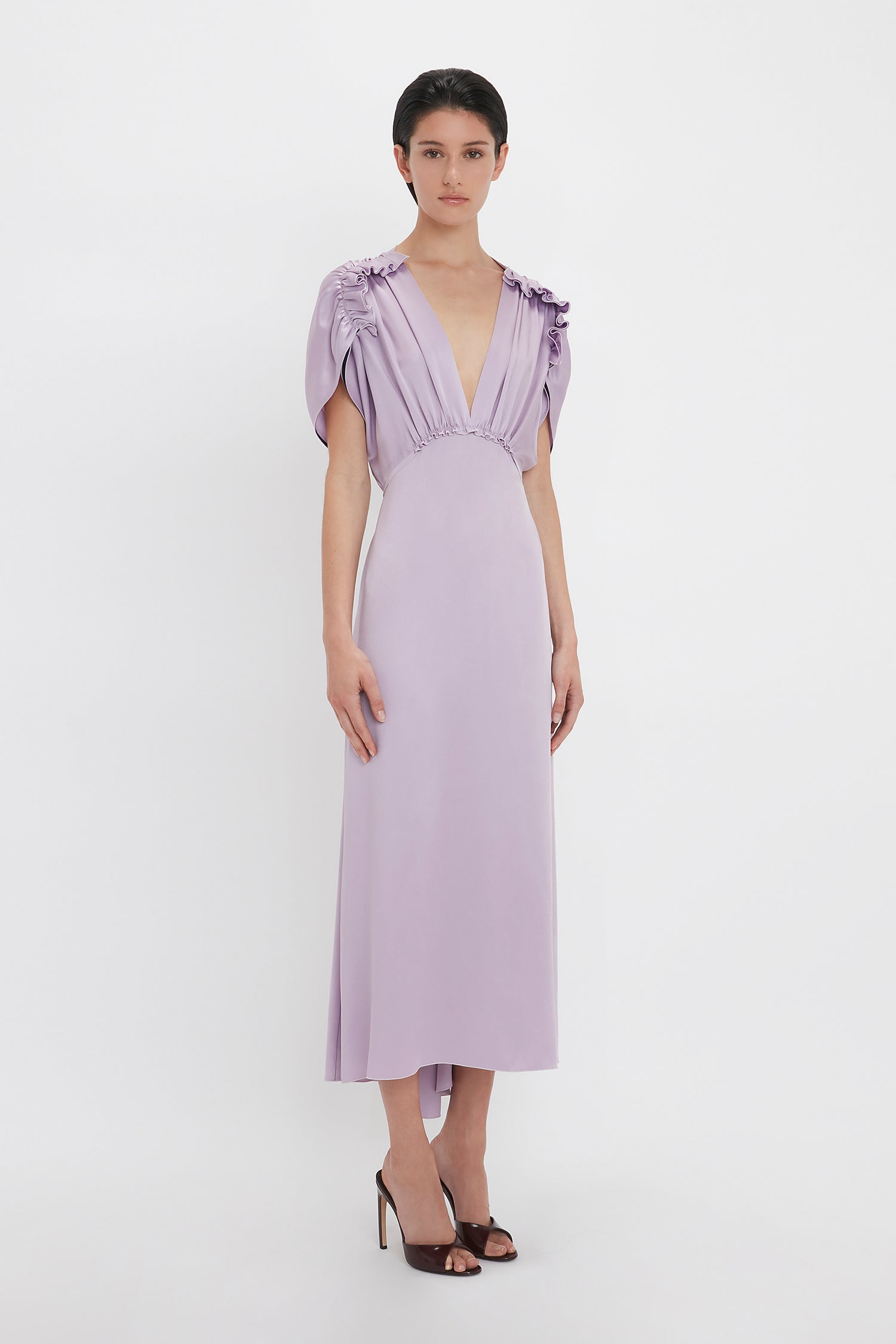 A person is wearing the Victoria Beckham V-Neck Ruffle Midi Dress In Petunia with short, draped sleeves and a deep V-neckline. Reminiscent of the Victoria Beckham brand’s sophisticated touch, they are standing against a plain, white background.