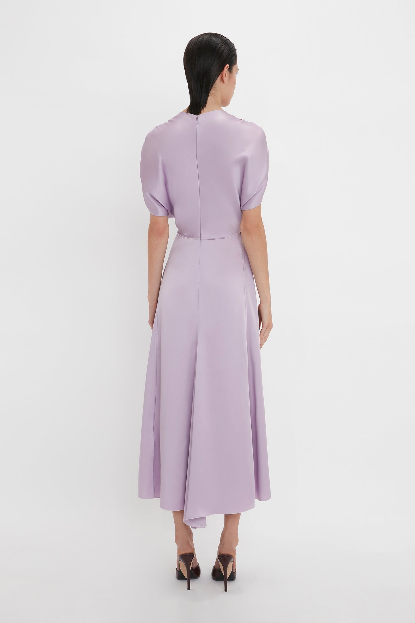 A woman with dark hair wearing an elegant light purple, short-sleeved dress and black heels is standing with her back to the camera on a plain white background, showcasing the timeless style of the V-Neck Ruffle Midi Dress In Petunia by Victoria Beckham.