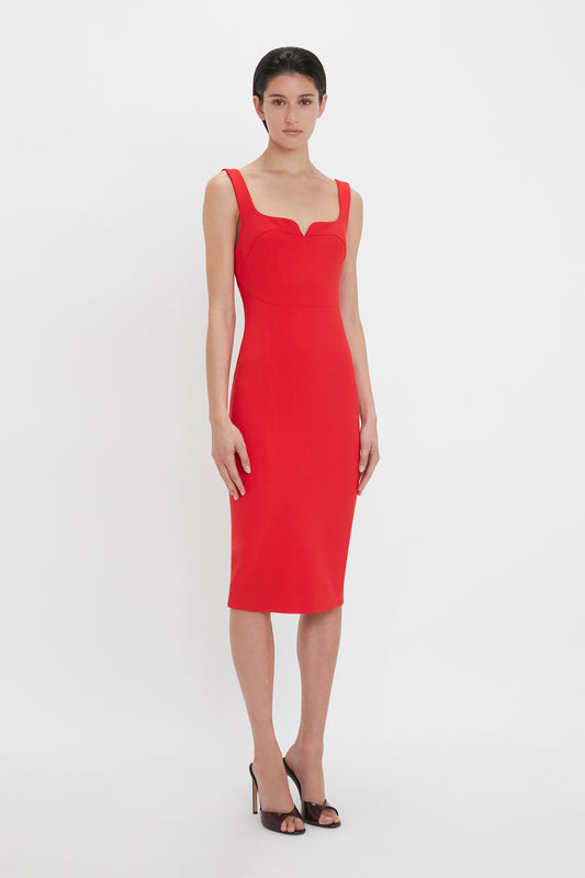 A person stands against a plain white background, wearing the Sleeveless Fitted T-Shirt Dress In Bright Red by Victoria Beckham, paired with black high-heeled shoes.