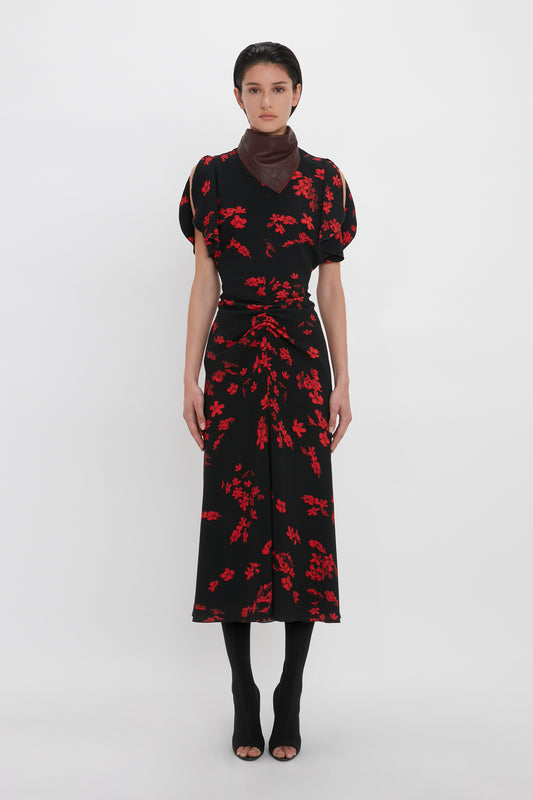 A person stands against a white background, wearing the stunning Gathered Waist Midi Dress In Sci-Fi Black Floral by Victoria Beckham. The brown neck scarf adds a touch of elegance, and their black open-toe shoes complete the chic look.