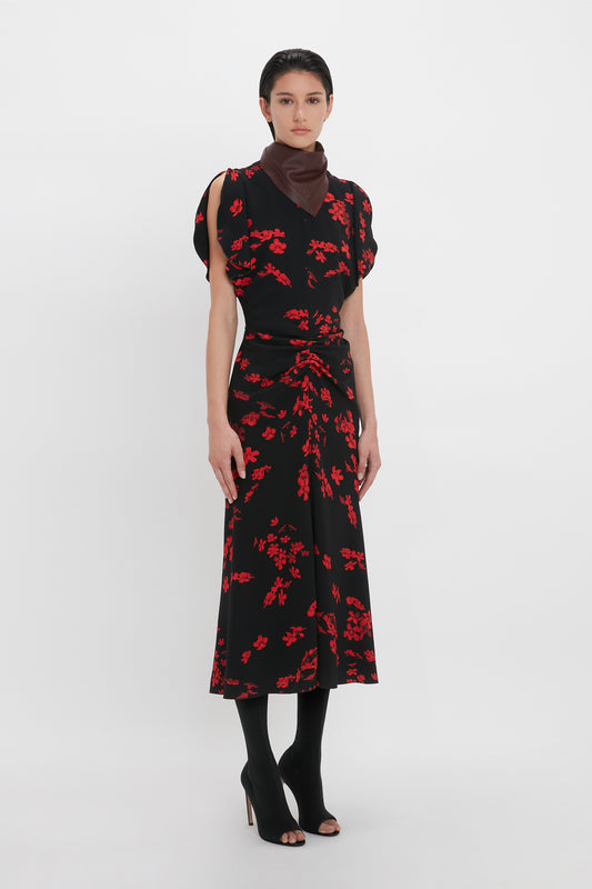 A person dressed in a Gathered Waist Midi Dress In Sci-Fi Black Floral by Victoria Beckham, a dark scarf, black tights, and open-toe heels stands against a plain background.