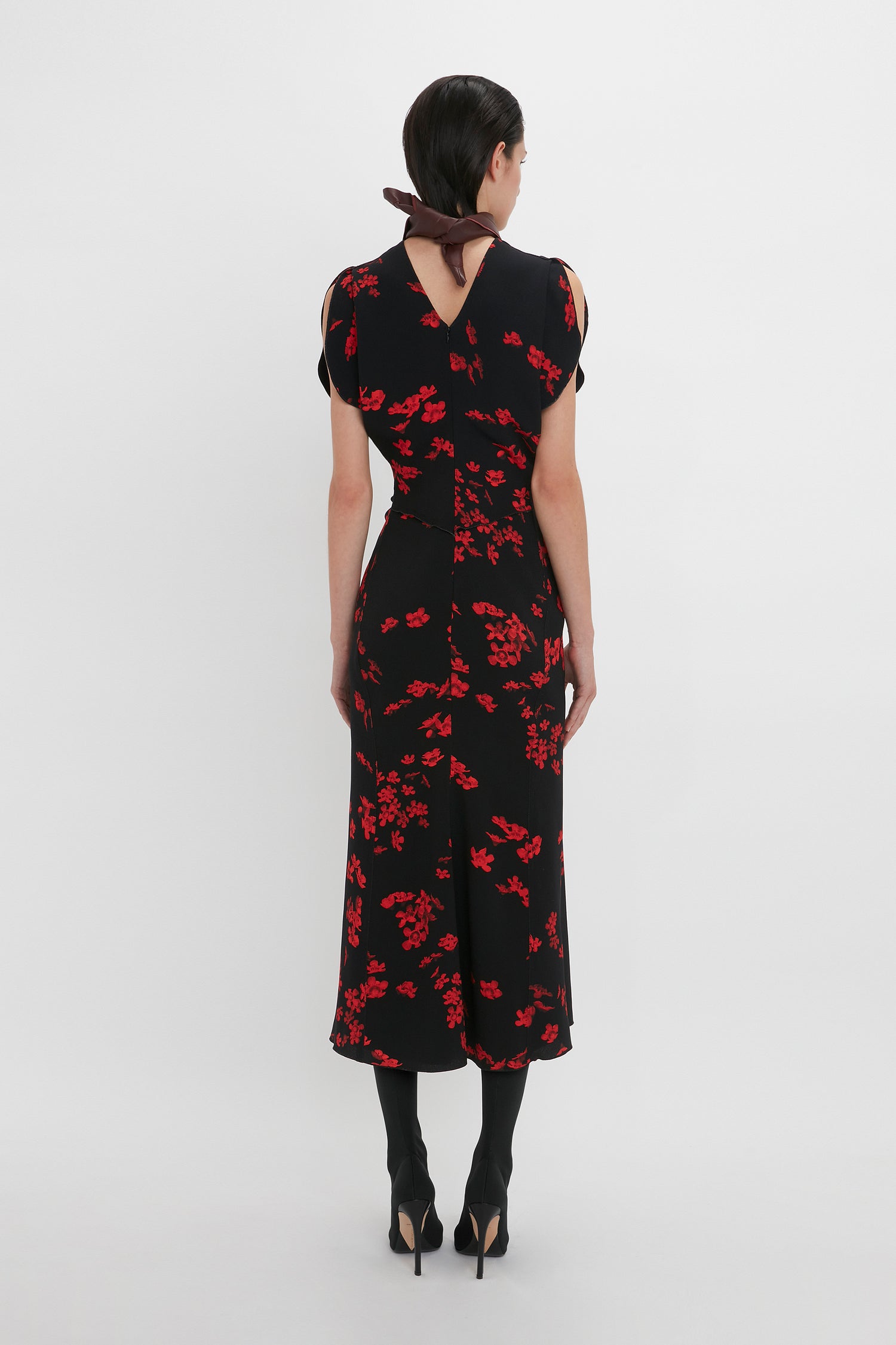 A person with dark hair in a ponytail is wearing a Gathered Waist Midi Dress In Sci-Fi Black Floral by Victoria Beckham featuring red floral patterns and a fit-and-flare silhouette, paired with black tights and high heels, standing with their back facing the camera against a plain white background.