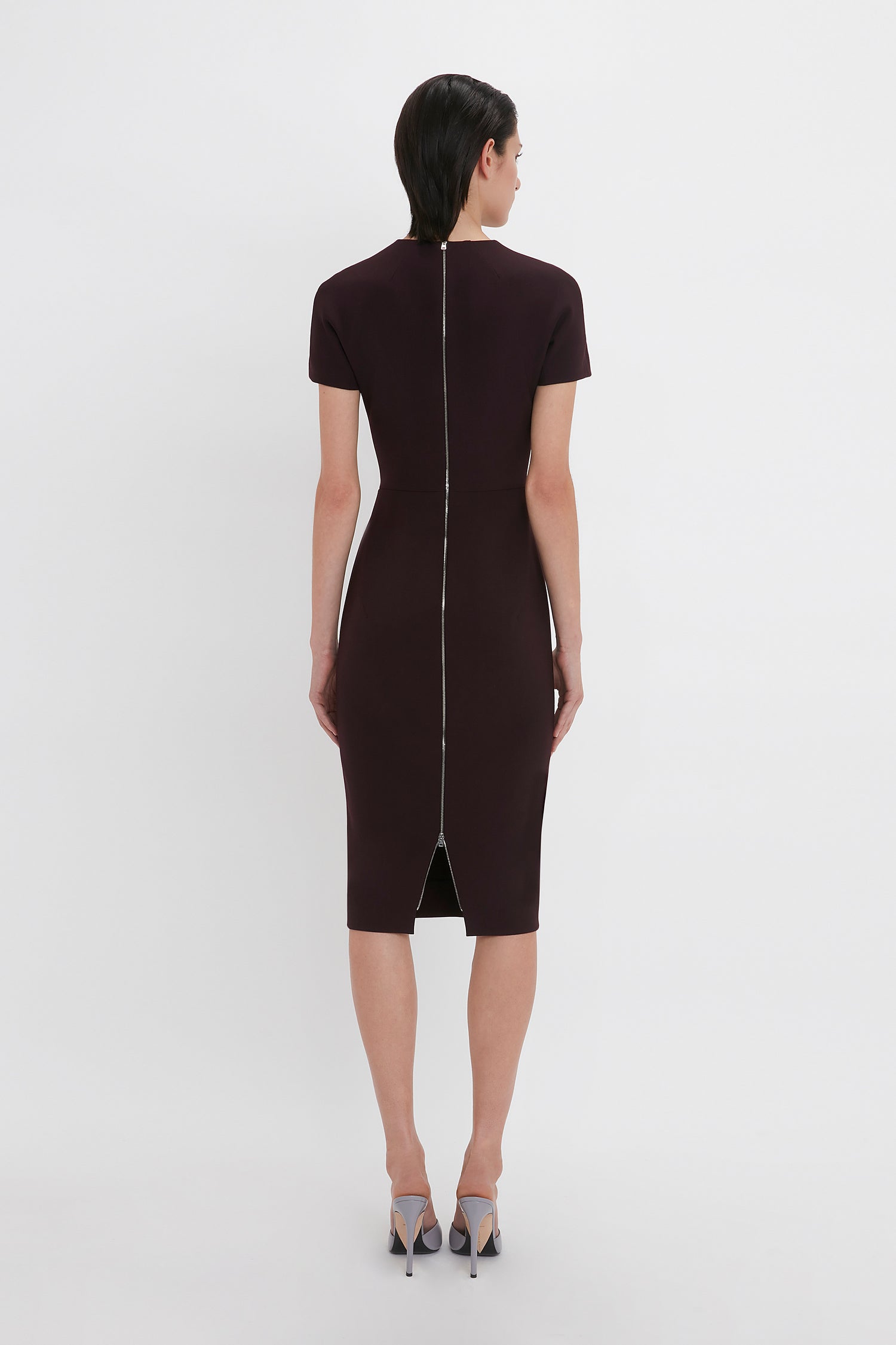 A person with short dark hair is wearing a Fitted T-Shirt Dress In Deep Mahogany, Victoria Beckham with a zipper running down the back, standing against a plain white background.