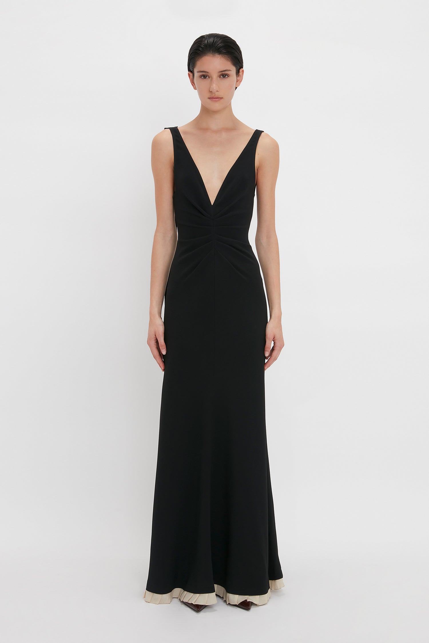 A person stands against a white background, wearing a sleeveless, floor-length black gown with a deep V-neckline and pleated details. The **V-Neck Gathered Waist Floor-Length Gown In Black** by **Victoria Beckham** features a gathered waist and a contrasting light-colored hem, adding an elegant touch.