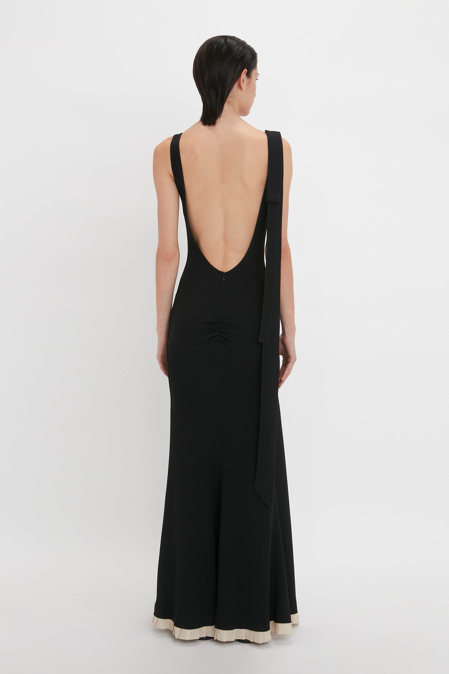 A person with short dark hair wearing a sleeveless, backless Victoria Beckham V-Neck Gathered Waist Floor-Length Gown In Black stands against a plain white background, facing away from the camera.