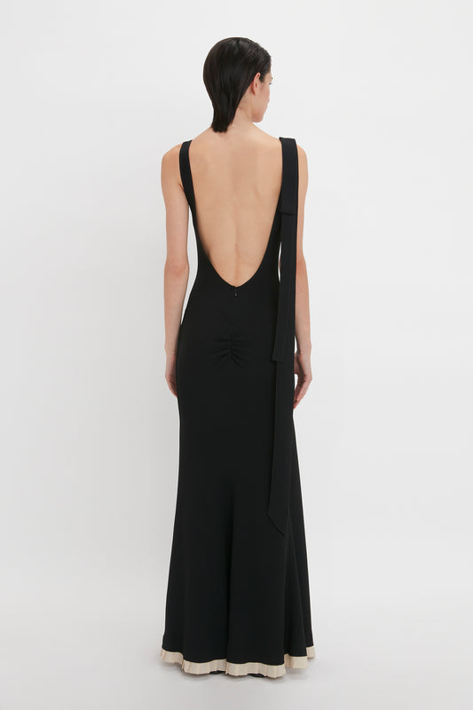 A person with short dark hair wearing a sleeveless, backless Victoria Beckham V-Neck Gathered Waist Floor-Length Gown In Black stands against a plain white background, facing away from the camera.