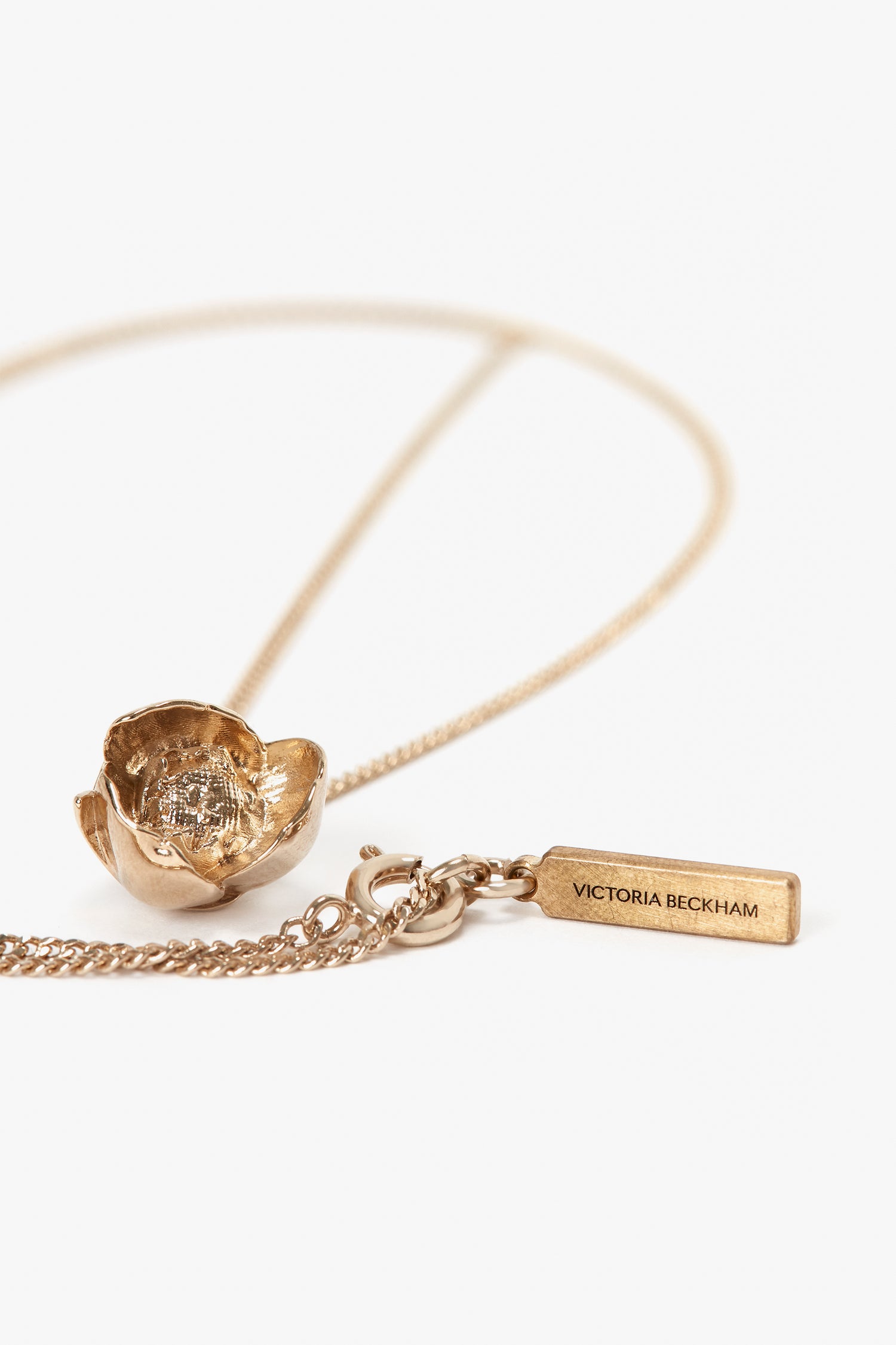 An **Exclusive Camellia Flower Necklace In Gold** featuring a pendant resembling a Camellia Flower and a rectangular tag inscribed with "**Victoria Beckham**." The chain, handcrafted in Italy, has a lobster clasp for closure.