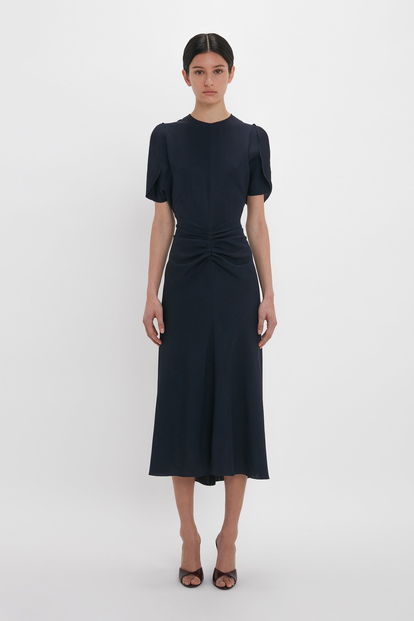 A person stands against a plain white background, wearing the Victoria Beckham Gathered Waist Midi Dress In Midnight with short sleeves and gathered detailing at the waist, paired with brown open-toe heels.