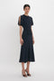 A person stands against a white background wearing the Victoria Beckham Gathered Waist Midi Dress In Midnight and black open-toe heels. The classic fit-and-flare design accentuates the gathered waist, adding elegance to the outfit.