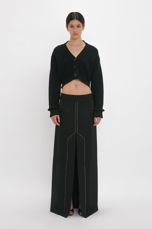 A person stands against a white background, dressed in a black cropped cardigan with large buttons and a Deconstructed Floor-Length Skirt In Black by Victoria Beckham, featuring front seams, creating an elongating silhouette.