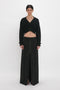 A person stands against a white background, dressed in a black cropped cardigan with large buttons and a Deconstructed Floor-Length Skirt In Black by Victoria Beckham, featuring front seams, creating an elongating silhouette.