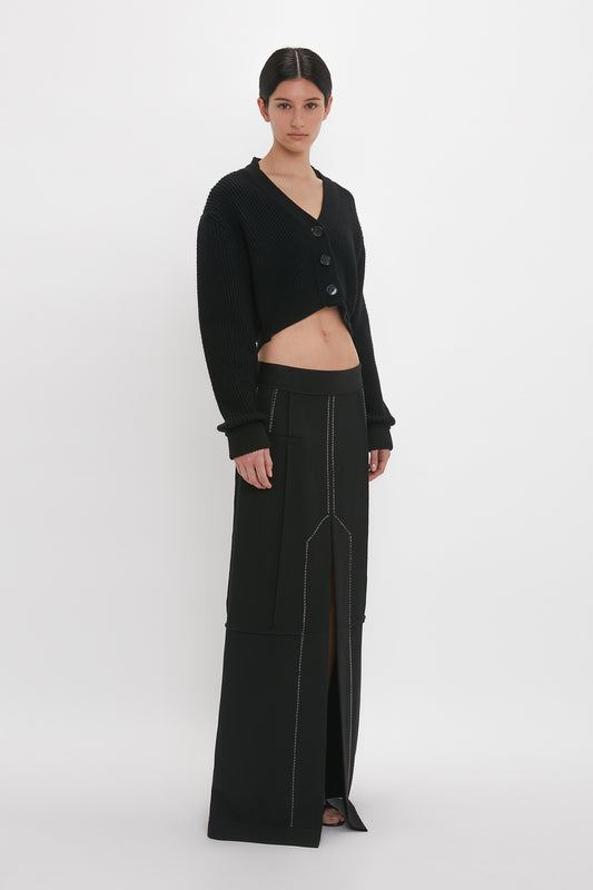A person is wearing a black cropped sweater and a floor-grazing Deconstructed Floor-Length Skirt In Black by Victoria Beckham with a center slit, creating an elongating silhouette. They are standing against a plain white background.