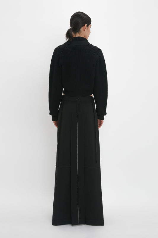 A person with dark hair in a low bun is standing facing away, wearing a black long-sleeve sweater and a Deconstructed Floor-Length Skirt In Black by Victoria Beckham that creates an elongating silhouette against a plain white background.