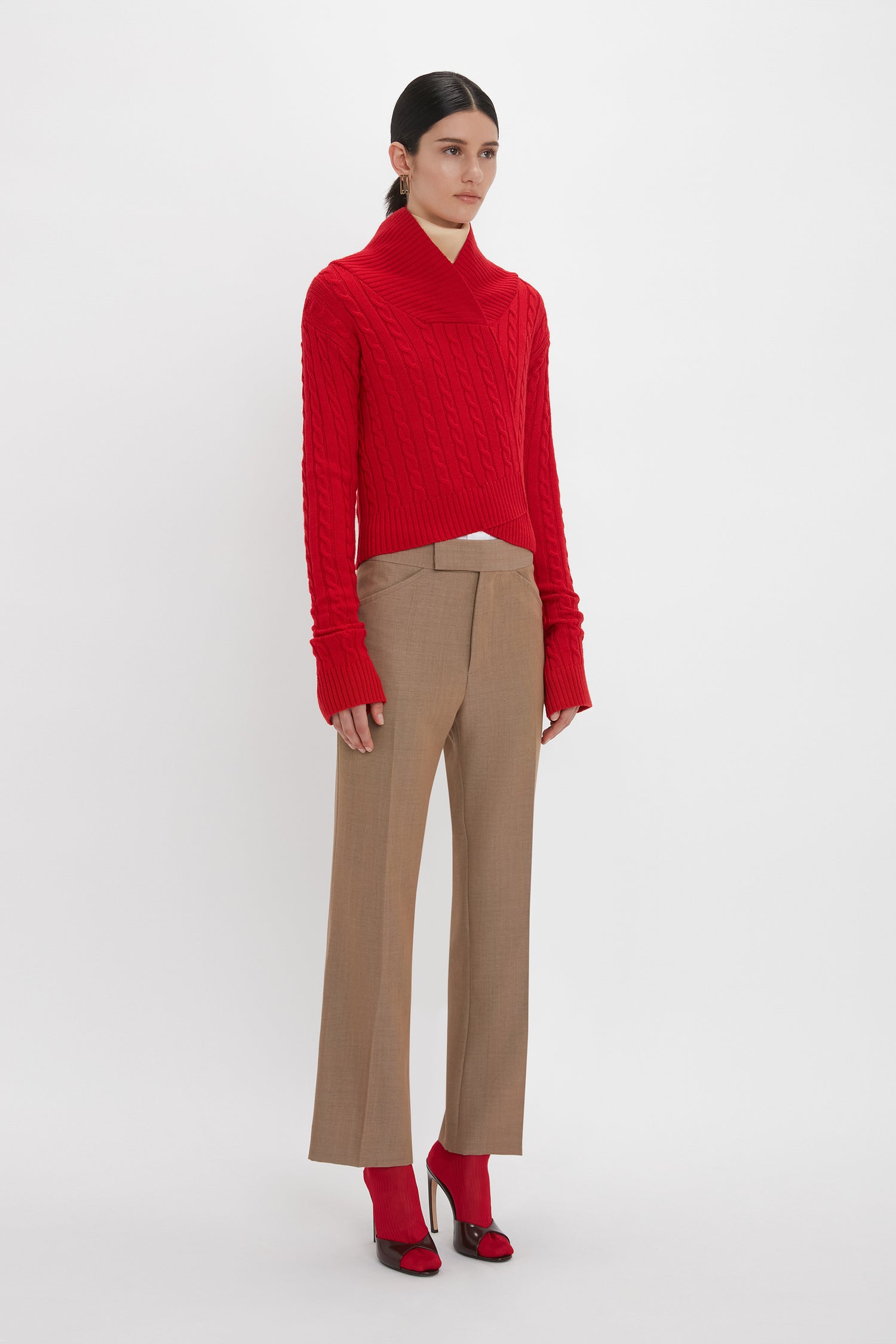 A person is standing against a plain background, wearing a red Victoria Beckham Wrap Detail Jumper In Red, tan trousers, and red high heels. They are looking to the side with a neutral expression, showcasing an elegant Victoria Beckham inspired style.